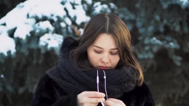 Jovem mulher usando fones de ouvido brancos ao ar livre no inverno. Jovem mulher atraente de casaco preto vestindo fones de ouvido de corda puxada branca no fundo do abeto no inverno — Vídeo de Stock