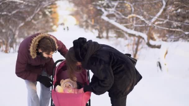 Ungt par promenader med bebis i barnvagn på vintern. Ung gift par ser in vagnen till happy baby medan promenader. Glad ung familj på promenad i park i vinter — Stockvideo