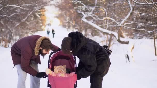 Молода пара гуляє з дитиною в колясці взимку. Молода подружня пара дивиться на коляску щасливою дитиною під час ходьби. Щаслива молода сім'я на прогулянці в парку взимку — стокове відео