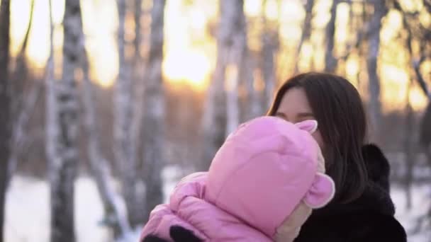 在户外抱着穿着连身裤的婴儿的女人。特写镜头的年轻女子与婴儿在粉红色的连身裤背景下的冬季小树林与日落 — 图库视频影像