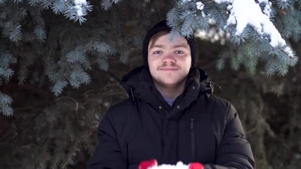 Ung man stående på bakgrund av Gran på vintern. Ung vacker man står under spruce och vacker kastar snö — Stockvideo