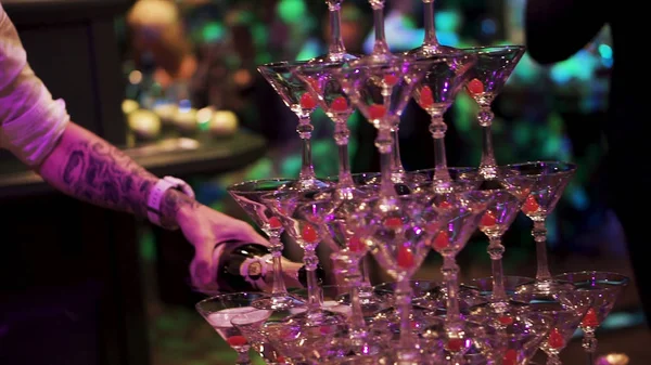 The waiter filled champagne fountain pyramid of glasses. Bottle of champagne being poured into a glass pyramid. People watching the waiter creates a pyramid of champagne glasses