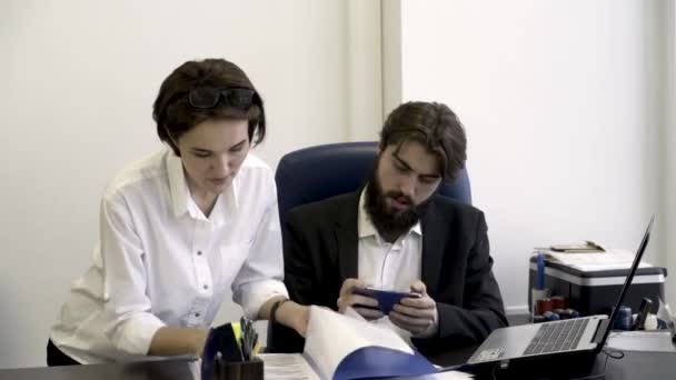 Женщина, офисный работник показывает важные документы своему бородатому боссу, который занят игрой в смартфоны в офисе. Зависимый бизнесмен не слушает своего секретаря с бумагами . — стоковое видео