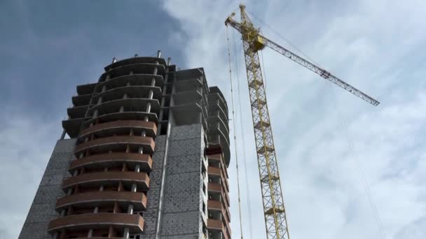 Construção de um edifício alto com um guindaste contra um céu azul com nuvens. Cena. Vista inferior. edifício cinzento de vários andares no processo de construção com janelas vazias com lixo de construção — Vídeo de Stock