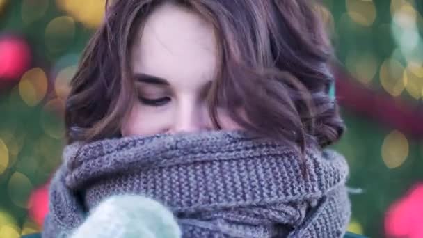 Hermosa joven con gran bufanda sonriendo al aire libre en invierno, fondo claro de Navidad. Retrato de invierno al aire libre de una niña sonriente en una bufanda de punto cerca del árbol de Navidad — Vídeo de stock