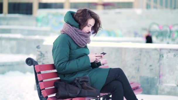 Mujer feliz enviando mensajes de texto en un teléfono inteligente mientras se sienta en un banco en la ciudad de invierno contra bloques de piedra con fondo de graffiti. Hermosa morena navegando en su teléfono móvil mientras descansa en un banco . — Vídeo de stock