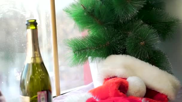 Sombrero de Santa Claus en el alféizar blanco contra la ventana y ramas de abeto artificial. Sombrero rojo, botella abierta de vino espumoso, ramas artificiales del árbol de Navidad, vacaciones de invierno y concepto de fiesta . — Vídeos de Stock