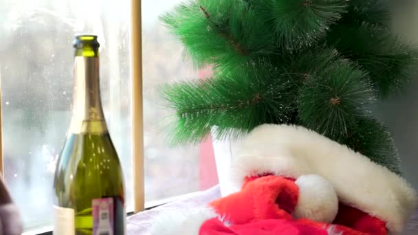Cappello Babbo Natale sul davanzale bianco contro la finestra e rami di abete rosso artificiale. Cappello rosso, bottiglia aperta di spumante, rami artificiali di albero di Natale, vacanze invernali e concetto di festa . — Video Stock