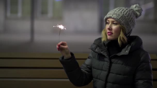 Smiling, beautiful girl in knitted hat and down jacket in the street at night with sparkler, celebrating New year, Merry Christmas. Young, pretty woman holding bengal fire on grey street background. — Stock Video