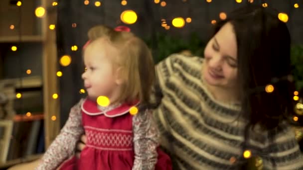Young, attractive, smiling woman mother with a cheerful, sweet child in beautiful New Year studio behind the yellow lights of garlands. Mom and baby girl in decorated room, Christmas Eve. — Stock Video
