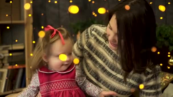 Jeune, jolie et souriante mère avec un enfant gai et doux dans le beau studio du Nouvel An derrière les lumières jaunes des guirlandes. Maman chatouille bébé fille dans la chambre décorée, veille de Noël . — Video