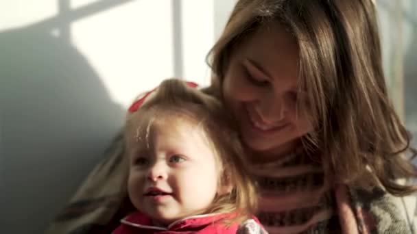 Vacker flicka med söt knubbig kinder leende i famnen mor. Härliga barn och hennes mamma mot fönstret njuta tillbringa tid tillsammans. — Stockvideo