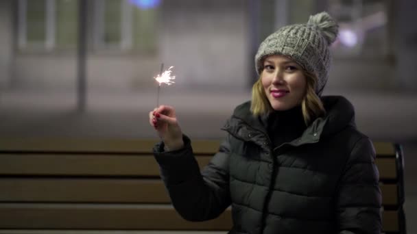 Jovem mulher sentada no parque com faíscas. Menina bonita sentada em um banco com sparklers — Vídeo de Stock