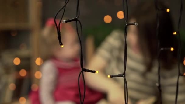 Jonge moeder met dochter lezing boekje thuis. Moeder en dochtertje spelen op de achtergrond van de slingers. Vervagen — Stockvideo