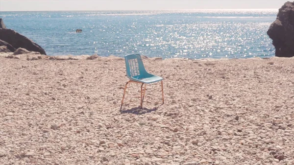 Chair on beach against sea. Stock. Old lonely chair stands on stone beach against blue beautiful sea and clear sky. Beach holiday by sea alone