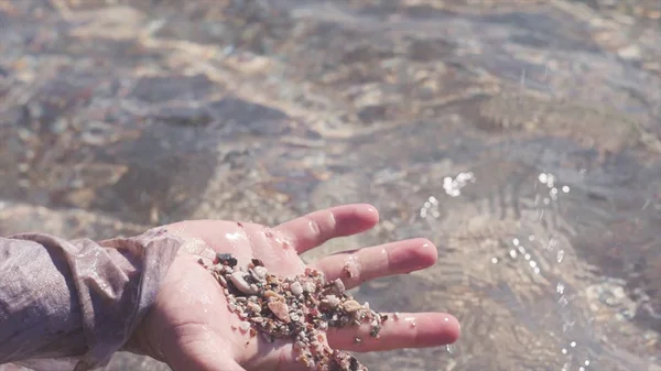Close-up hand takes out of water handful of sand. Stock. Close-up man examines sand stones from bottom of clear water. Beautiful shiny clear water in sun. Summer holidays by water