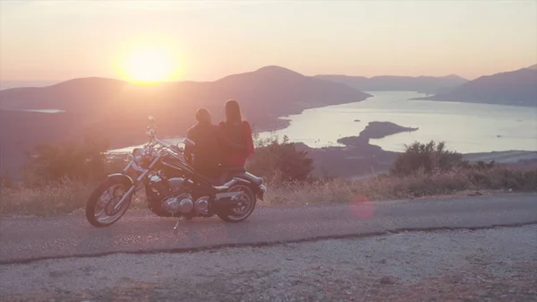 Widok z tyłu romantyczna para z motocykla o zachodzie słońca. Magazynie. Romantyczny daty motocyklista i kobieta w czerwonej sukience na tle zachodu słońca i górskich scenerii z rzeki — Zdjęcie stockowe