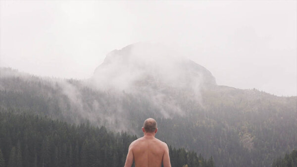 View from back of man on background mountains and lake. Stock. Man with naked torso in jeans stands in rain and looks at beautiful mountain landscape with green forest by lake
