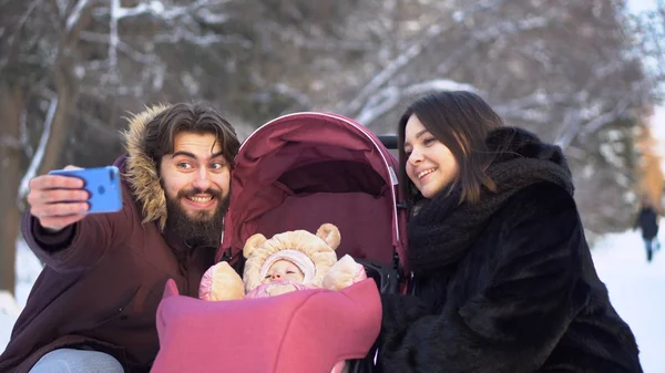 Pareja con bebé tomar selfie a pie. Joven pareja casada con el bebé en el cochecito tomar selfie en caminar en invierno. Moderna familia feliz joven —  Fotos de Stock