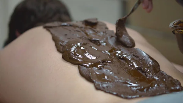Massage with mud on back. Close-up massage therapist of Spa salon smears brown mixture on male back. Spa care, relaxation and treatment