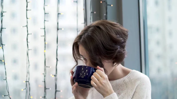 Красивая женщина пьет чай и смотрит в окно с гирляндами. Зимний теплый комфорт. Женщина смотрит в окно в ожидании прибытия любимого человека — стоковое фото
