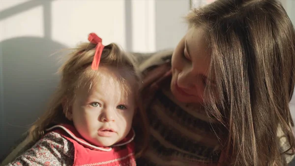 Jonge vrouw met een schattig klein meisje in de buurt van venster thuis. Jonge moeder en dochter zitten bij het raam van woning — Stockfoto