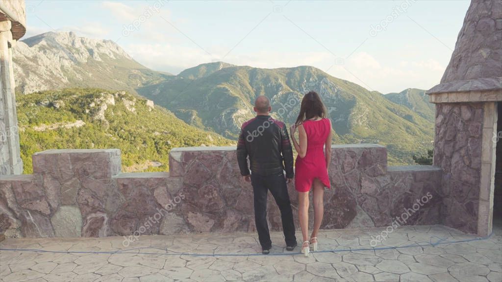Back view of couple looking from fortress to mountains. Stock. Date of man in leather jacket with sexy woman in red dress against mountains. Lovers together observing romantic view