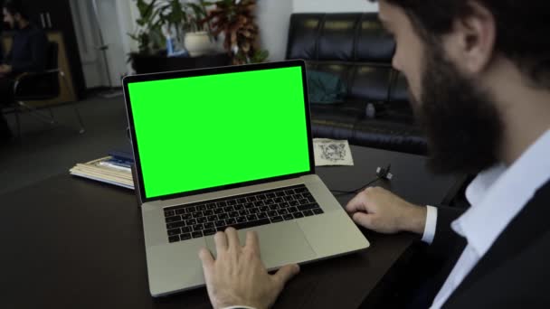 Um jovem usando um laptop com uma tela verde dentro de casa. Um plano de perto. Homem barbudo trabalhando no escritório para um laptop com tela verde — Vídeo de Stock