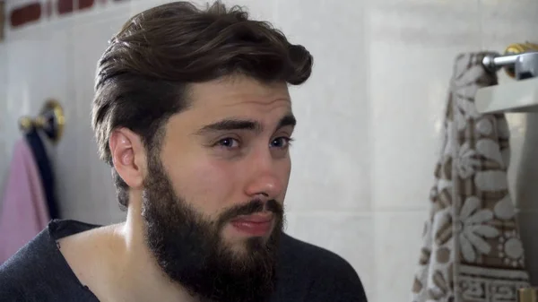 Portrait of young teenager man looking at himself in a home bathroom mirror doing his hair and getting ready in the morning, home interior. Handsome unshaven man looking in mirror — Stock Photo, Image