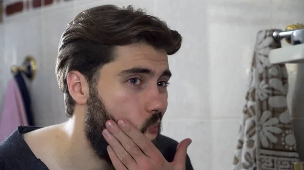 Портрет молодого чоловіка-підлітка, який дивиться на себе в дзеркало домашньої ванної кімнати, робить волосся і готується вранці, домашній інтер'єр. Красивий беззахисний чоловік дивиться у дзеркало — стокове фото
