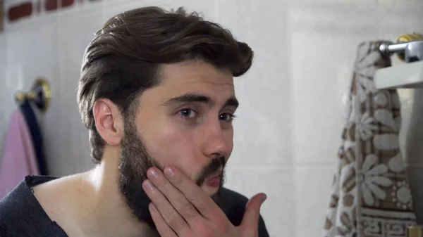 Retrato de jovem adolescente olhando para si mesmo em um espelho do banheiro em casa fazendo seu cabelo e se preparando pela manhã, interior da casa. Bonito homem raspado olhando no espelho — Fotografia de Stock