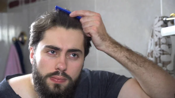 Un hombre en el baño mirándose en un espejo y arreglándose el pelo. Retrato de un hombre guapo cepillándose el pelo en el baño. Reflexión en el espejo de un hombre guapo peinando el cabello — Foto de Stock