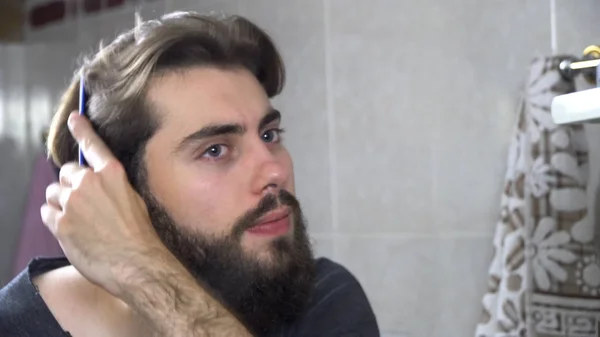 Um homem na casa de banho a olhar para o espelho e a arranjar o cabelo. Retrato de um homem bonito a escovar o cabelo na casa de banho. Reflexão no espelho de homem bonito styling cabelo — Fotografia de Stock