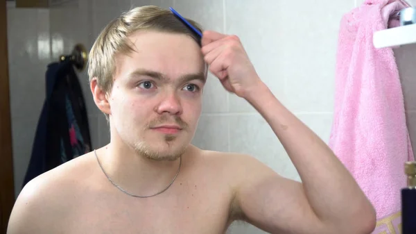 Um homem na casa de banho a olhar para o espelho e a arranjar o cabelo. Retrato de um homem bonito a escovar o cabelo na casa de banho. Reflexão no espelho de homem bonito styling cabelo — Fotografia de Stock