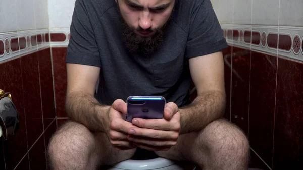 Nahaufnahme eines jungen kaukasischen Mannes, der auf der Toilette sitzt und mit seinem Smartphone eine Nachricht sendet. Nahaufnahme eines jungen kaukasischen Mannes mit seinem Smartphone im Badezimmer — Stockfoto