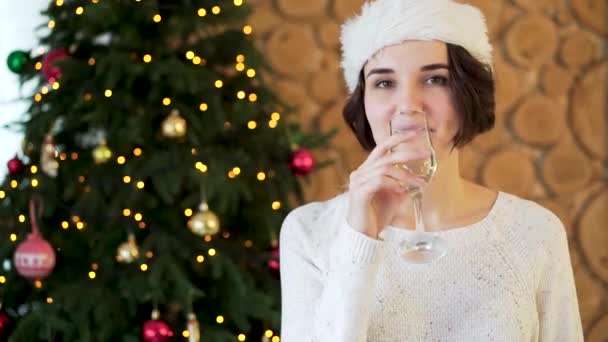 穿着毛衣和长袜的女孩坐在圣诞树的背景上, 拿着一杯香槟酒。女孩手里拿着一杯香槟酒庆祝圣诞节. — 图库视频影像