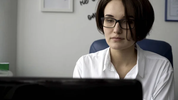 Da vicino per una giovane donna in camicia bianca che guarda il computer portatile, risponde al telefono e sorride sullo sfondo bianco della parete dell'ufficio. Imprenditrice che lavora davanti allo schermo del computer nel suo posto di lavoro . — Foto Stock