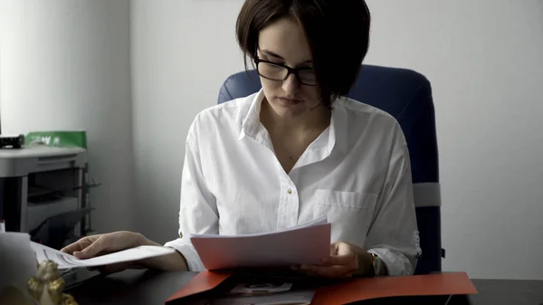 Красивая женщина с короткими темными волосами в очках раскладывает документы из красной папки на белом фоне офисной стены. Концентрированная офисная леди в белой рубашке, работающая с бумагами, бизнес-концепция . — стоковое фото