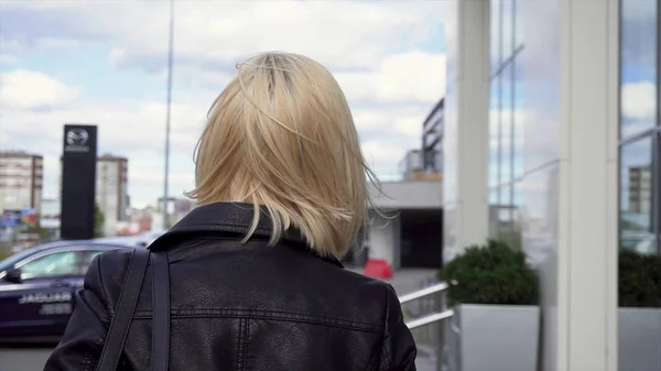 Vista trasera de una rubia en una chaqueta de cuero negro caminando por la calle de la ciudad. Acciones. Mujer joven y rubia caminando por las calles de la ciudad con una bolsa sobre su hombro . — Foto de Stock