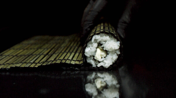 Процесс суши прокатки с черным бамбуком макису, изолированный на черном фоне. Руки в черных перчатках катятся суши с черным бамбуковым ковриком, концепция приготовления
.
