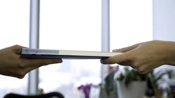 Passing file folder from hands to hands on fuzzy, office window background, teamwork concept. Two young hands of colleagues give each other blue folder.