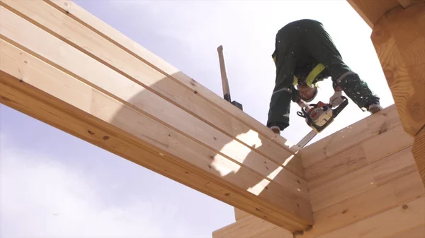 Vue du bas d'un homme en uniforme debout sur des poutres en bois, à l'aide d'une tronçonneuse à sciure de bois volant partout. Constructeur se tient sur le cadre en bois de la maison avec une scie et des copeaux de bois contre le ciel . — Photo