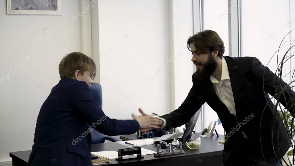 Cheerful male friends in business suits greeting each other with funny ritual greeting dance on office furniture background. Two happy colleagues and pals greet each other by special handshaking