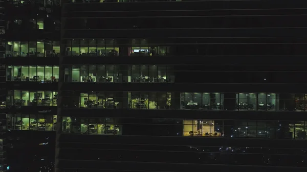 Vue frontale de la façade nocturne du bâtiment avec beaucoup de fenêtres éclairées. Fusillade. Façade du bâtiment multi-étages de verre et d'acier, bureaux et travailleurs à l'intérieur, fond de la vie nocturne de la ville — Photo