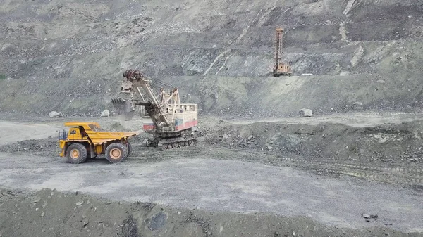 Excavator and dump truck while loading stone ore in a grey quarry, mining industry. Stock. Heavy mining excavator loads rock ore into a dump-body large mining truck, quarry equipment.