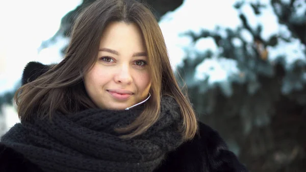 Närbild av unga, attraktiva tjejen i en päls och stickad halsduk tittar rakt i en vinter park, gatumode. Porträtt av vacker kvinna i vinterkläder på spruce grenar bakgrund. — Stockfoto