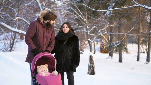 Familia joven y feliz caminando en un parque de invierno, mamá, papá y bebé en un cochecito rosa. Hombre señalando el dedo y los padres mirando a la izquierda en el fondo de los árboles de invierno . —  Fotos de Stock