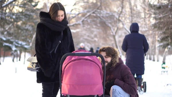 Familia joven y feliz caminando en un parque de invierno, mamá, papá y bebé en un cochecito. Padres sonrientes inclinados sobre un cochecito rosa y hablando con el bebé sobre el fondo de árboles nevados . — Foto de Stock