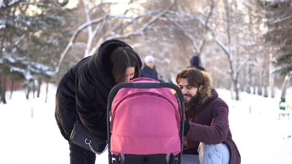 Familia joven y feliz caminando en un parque de invierno, mamá, papá y bebé en un cochecito. Padres sonrientes inclinados sobre un cochecito rosa y hablando con el bebé sobre el fondo de árboles nevados . —  Fotos de Stock