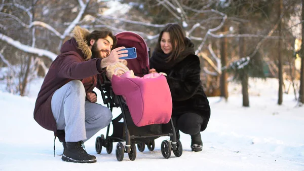Feliz, joven familia caminando, haciendo fotos en un parque de invierno, mamá, papá y el bebé en el cochecito. Hombre está haciendo selfie wirh su sonriente esposa y bebé en rosa cochecito en los árboles nevados fondo . —  Fotos de Stock